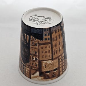 Puede incluir: Una taza de cer&aacute;mica con un dise&ntilde;o de paisaje urbano pintado a mano en tonos marr&oacute;n, beige y negro. La taza tiene un borde y una base blancos. El texto manuscrito en el borde interior dice "Ground Zero Studio View, 9/11, NYC".