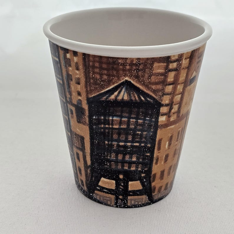 Puede incluir: Un vaso de caf&eacute; de papel desechable con un dise&ntilde;o de paisaje urbano. La taza presenta una ilustraci&oacute;n marr&oacute;n y beige de edificios y una torre de agua. El borde de la taza es blanco y el interior tambi&eacute;n es blanco.