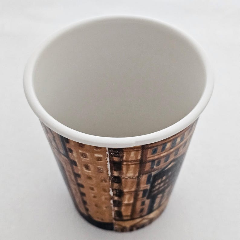 Puede incluir: Una taza de caf&eacute; desechable de papel con interior y borde blancos. El exterior presenta un dise&ntilde;o arquitect&oacute;nico marr&oacute;n y negro. La taza est&aacute; vac&iacute;a y se encuentra sobre una superficie blanca.