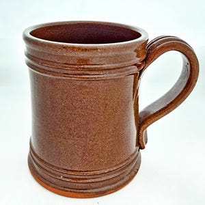 May include: A brown ceramic mug with a large handle. The mug has a cylindrical shape with horizontal ridges near the top and bottom. The interior of the mug is a darker shade of brown.
