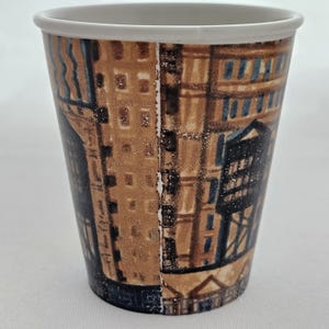 Puede incluir: Una taza de caf&eacute; de papel con un borde blanco y un dise&ntilde;o de paisaje urbano marr&oacute;n y azul. La taza presenta un patr&oacute;n repetitivo de edificios y elementos arquitect&oacute;nicos. La taza est&aacute; dise&ntilde;ada para bebidas calientes.