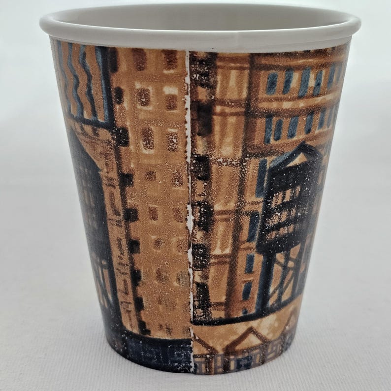 Puede incluir: Una taza de caf&eacute; de papel con un borde blanco y un dise&ntilde;o de paisaje urbano marr&oacute;n y azul. La taza presenta un patr&oacute;n repetitivo de edificios y elementos arquitect&oacute;nicos. La taza est&aacute; dise&ntilde;ada para bebidas calientes.
