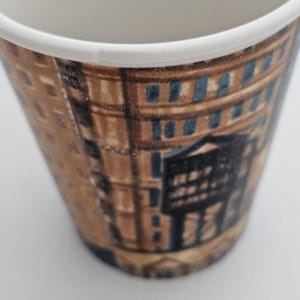 Puede incluir: Taza de caf&eacute; de papel con una detallada ilustraci&oacute;n de una fachada de edificio en tonos marrones, beige y azules. La taza tiene un borde blanco y una forma c&oacute;nica, ideal para bebidas calientes. El dise&ntilde;o evoca la arquitectura urbana.