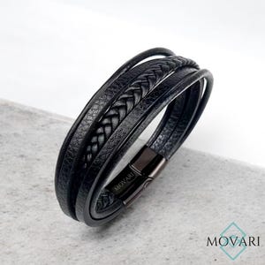 May include: Black leather bracelet with multiple strands, including a braided section. The bracelet has a magnetic clasp and the brand name "MOVARI" is visible. The bracelet is displayed on a light gray surface.