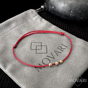 May include: Red string bracelet with three gold beads. The bracelet is on a gray velvet pouch with the word "MOVARI" printed on it.