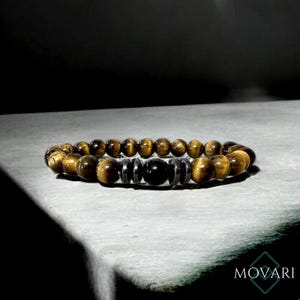 May include: A brown and gold beaded bracelet with a black bead in the center. The bracelet is made of tiger's eye and hematite beads.