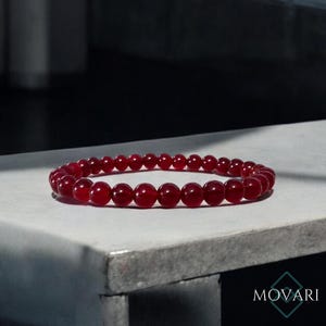 May include: A red gemstone bracelet made of round beads. The bracelet is on a gray surface.
