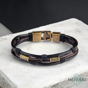 May include: A multi-strand leather bracelet in black and brown with antique brass accents. The bracelet features a clasp closure and decorative metal beads. The bracelet is displayed on a white surface.