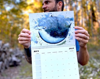 May include: A 2027 wall calendar featuring a watercolor illustration of a hammerhead shark swimming in front of a large, dark blue circular shape. The page is titled July 2027 and displays a grid of dates for the month with black text.