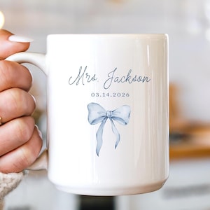 May include: White ceramic mug featuring a blue bow illustration, the text "Mrs. Jackson" and the date "03.14.2026". The mug has a large handle and a smooth, glossy finish. The design is simple and elegant.