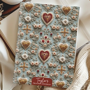 Puede incluir: Un diario de oración con una cubierta azul claro con un estampado floral con corazones dorados, cruces y detalles en rojo. El diario tiene el texto "Taylor's Prayer Journal" en la parte inferior.