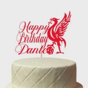 Puede incluir: Tarta blanca de dos pisos con un patrón de diamantes, coronada con un adorno rojo. El adorno lleva el texto "Happy Birthday Dante" y un emblema de pájaro rojo.