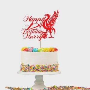 Puede incluir: Una tarta de cumpleaños blanca con chispas y glaseado coloridos, coronada con un adorno rojo que dice "Happy 12th Birthday Harry" y presenta un emblema de pájaro estilizado. La tarta está sobre un pedestal blanco.