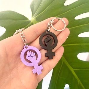 Könnte beinhalten: Zwei Schlüsselanhänger mit dem feministischen Symbol und einem erhobenen Faust-Design. Einer ist lavendelfarben, der andere schwarz. Beide haben silberfarbene Schlüsselringe und Ketten. Der Hintergrund ist ein großes grünes Blatt.