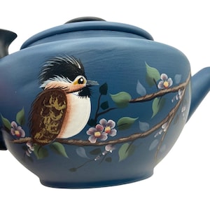 May include: A blue ceramic teapot with a black handle and lid. The teapot is decorated with a hand-painted bird perched on a branch with flowers and leaves. The bird has brown, white, and black feathers. The teapot has a capacity of approximately 4 cups.