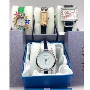 May include: A collection of women's watches, each displayed in individual boxes. The watches feature various designs, including floral accents, rectangular and square faces, and different band colors. One watch has the brand name "STRADA" on the watch face.