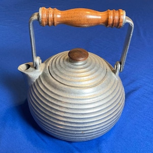 May include: A vintage silver-toned kettle with a wooden handle and lid knob. The kettle has a round body with horizontal ridged detailing. The handle is attached with metal brackets. The lid knob is a dark brown color.