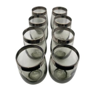 May include: Eight stemless wine glasses with a smoky gray hue and a silver rim. The glasses are arranged in two rows of four, showcasing their elegant design and shape. The clear glass allows the contents to be visible.