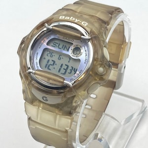 May include: A translucent brown Baby-G digital watch by Casio with a round case and matching resin strap. The watch face displays the text Baby-G, Casio, and digital readouts including the day SUN, the date 26-6-7, and the time 12:13:34.