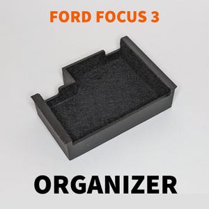 Puede incluir: Organizador negro para Ford Focus 3. El organizador rectangular tiene un interior texturizado y bordes elevados. El texto "FORD FOCUS 3" es naranja y "ORGANIZER" es negro.