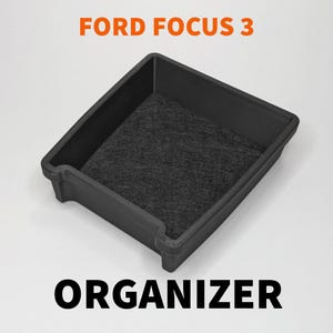 Puede incluir: Organizador negro para Ford Focus 3, con interior texturizado gris oscuro. El organizador es rectangular con lados ligeramente curvados. El texto "FORD FOCUS 3" y "ORGANIZER" está en naranja y negro.
