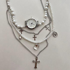 May include: A silver layered necklace with a pearl strand, a watch with a white face, and multiple cross charms. The necklace also features a crystal pendant.