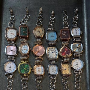 Puede incluir: Una colección de 12 relojes de pulsera plateados con diferentes caras y diseños de colores. Los relojes tienen correas de eslabones de cadena y están dispuestos en dos filas sobre una superficie gris.