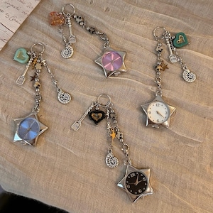 May include: Five silver-toned keychains with star-shaped pendants. Each keychain features a different design, including a clock face, a heart, and a gemstone. Additional charms include small stars, musical notes, and other decorative elements.