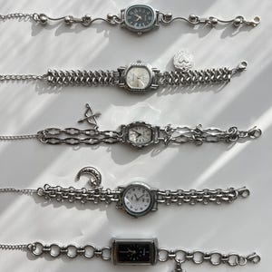 May include: A collection of silver-toned wristwatches with various chain and charm designs. The watch faces are in different shapes, including oval, square, and rectangular. Some watches have Roman numerals, while others have Arabic numerals. Charms include a cross, a crescent moon, and an arrow.
