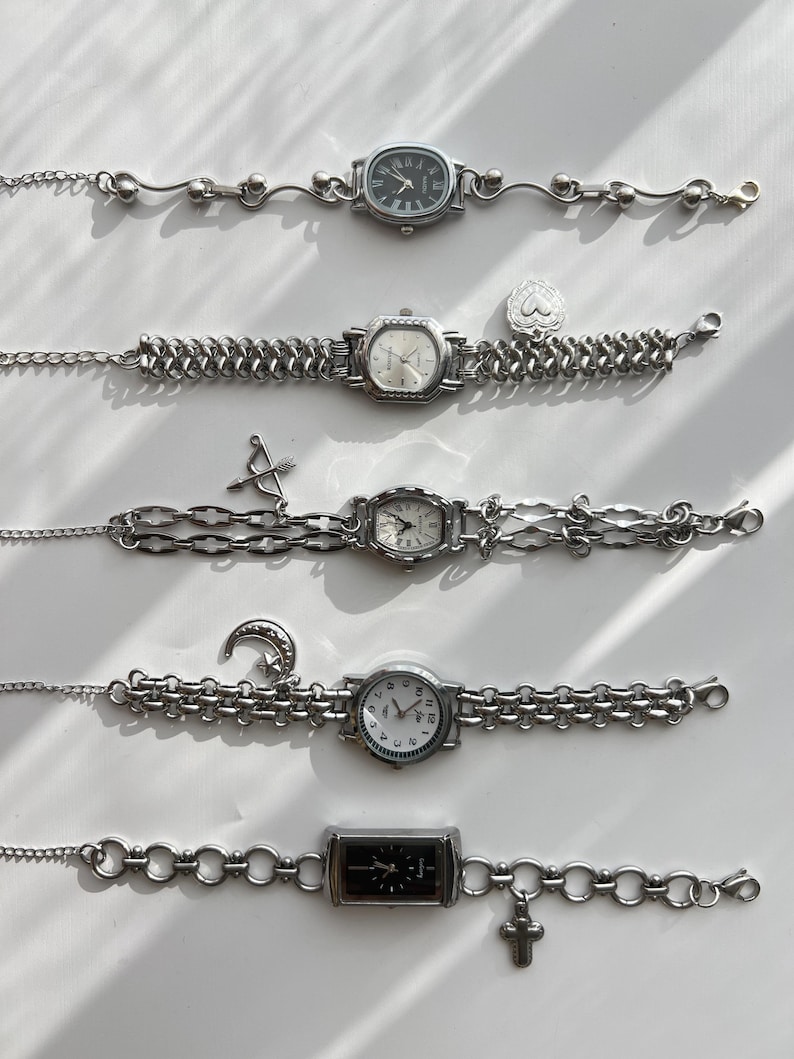 May include: A collection of silver-toned wristwatches with various chain and charm designs. The watch faces are in different shapes, including oval, square, and rectangular. Some watches have Roman numerals, while others have Arabic numerals. Charms include a cross, a crescent moon, and an arrow.