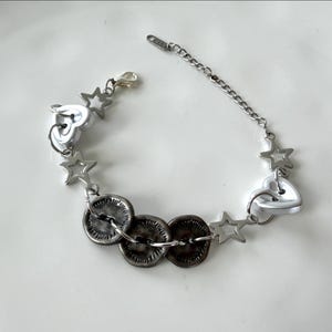 May include: Silver-toned bracelet featuring a mix of heart and star charms, along with three dark, textured circular elements. The bracelet has a chain extension for adjustable sizing and a lobster clasp closure.
