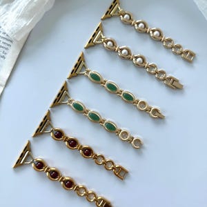 以下が含まれることがあります： 一列に並べられた6本のゴールドトーンの金属製時計バンド。各バンドは楕円形のリンクで構成されており、白い真珠、緑色の石、または濃い赤色の石がはめ込まれ、金属製のコネクタが付いています。