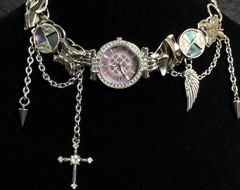 May include: A silver necklace with a watch, a cross pendant, a wing charm, and other decorative elements. The watch has a pink face and a silver band.
