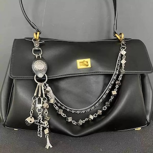 Pode incluir: Uma bolsa de mão de couro preto com uma alça de corrente prateada e um pingente prateado com dados e uma Torre Eiffel. A bolsa tem um fecho dourado e um pingente de flor prateado.