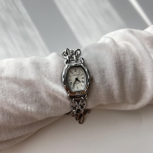 May include: A silver-toned wristwatch with an oval face and a white dial. The watch features Roman numerals and a chain-link band. The watch is displayed on a white fabric surface.