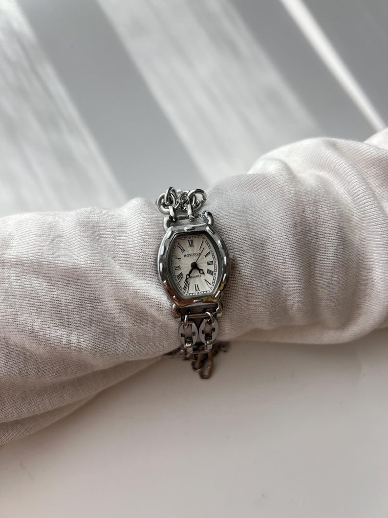 May include: A silver-toned wristwatch with an oval face and a white dial. The watch features Roman numerals and a chain-link band. The watch is displayed on a white fabric surface.