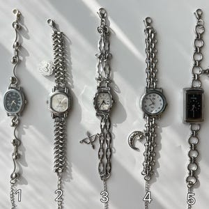 May include: Five silver-toned vintage wristwatches with various chain styles and watch faces. Each watch has a unique design, including oval, square, and round faces. Some feature decorative charms like a heart, a crescent moon, and a bow and arrow.