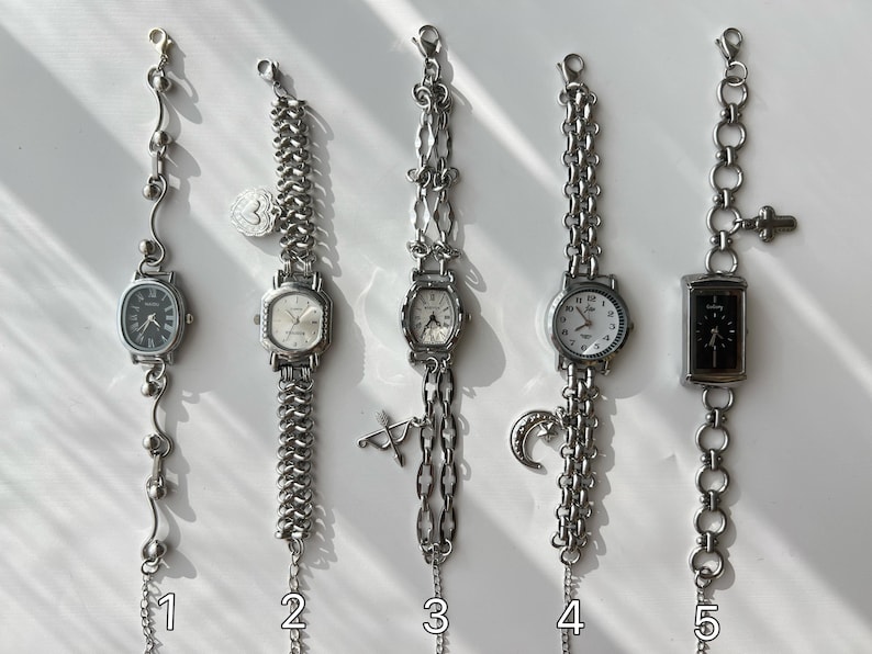 May include: Five silver-toned vintage wristwatches with various chain styles and watch faces. Each watch has a unique design, including oval, square, and round faces. Some feature decorative charms like a heart, a crescent moon, and a bow and arrow.