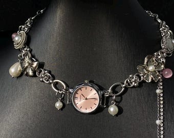 May include: A silver chain necklace with a pink watch face and pearl accents. The necklace features a bow, flowers, and other decorative charms.
