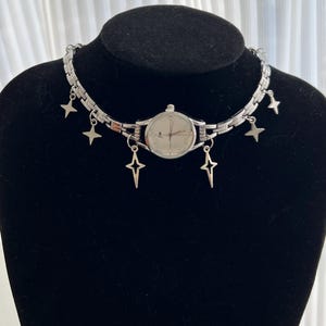 Op de afbeelding: Een zilveren horloge met een witte wijzerplaat en zilveren wijzers is bevestigd aan een zilveren ketting met stervormige bedels. Het horloge bevindt zich op een zwart manikin.