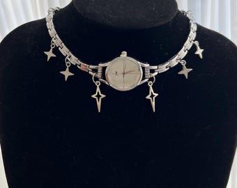 May include: A silver watch with a white face and silver hands is attached to a silver chain with star-shaped charms. The watch is on a black mannequin.