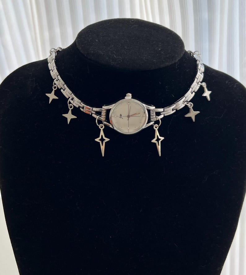 Op de afbeelding: Een zilveren horloge met een witte wijzerplaat en zilveren wijzers is bevestigd aan een zilveren ketting met stervormige bedels. Het horloge bevindt zich op een zwart manikin.