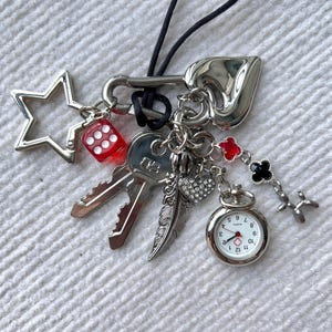 May include: A silver charm necklace featuring a selection of charms, including a star, a dice, a key, a feather, a heart, a pocket watch, and a clover. The charms are attached to a black cord.