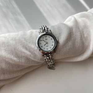 May include: A silver-toned quartz watch with a white face and black numerals. The watch has a linked band and the brand name "jew" is printed on the face. The watch is resting on a white fabric.