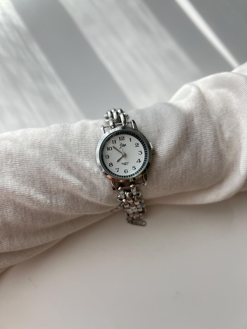 May include: A silver-toned quartz watch with a white face and black numerals. The watch has a linked band and the brand name "jew" is printed on the face. The watch is resting on a white fabric.