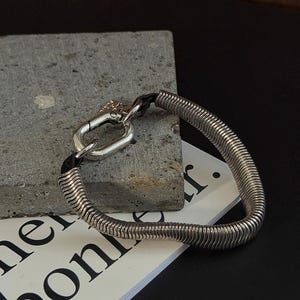 May include: A silver bracelet with a distinctive design. It has a coiled, spring-like band and a clasp. The bracelet is on a grey stone and a white paper with black text. The bracelet's design is eye-catching.