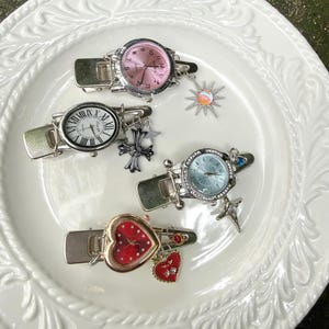 May include: Four decorative hair clips with watch faces. The clips have silver metal clasps and various watch faces in pink, white, and blue. Each clip has a charm, including a sun, cross, star, and heart. The clips are on a white, decorative plate.