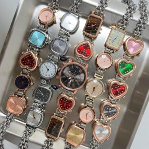 Puede incluir: Una colección de 17 relojes de pulsera de tono plateado y dorado con diferentes caras y correas de colores. Los relojes tienen caras cuadradas, rectangulares y en forma de corazón. Algunos de los relojes tienen marcas de números romanos en la cara.