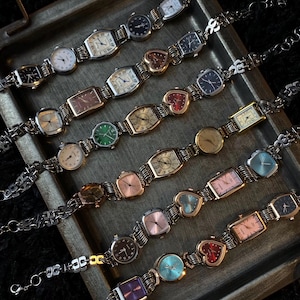Puede incluir: Una colección de relojes de pulsera de tono plateado y dorado con diferentes caras de colores y diseños en forma de corazón. Los relojes están dispuestos en una bandeja plateada con un fondo negro.