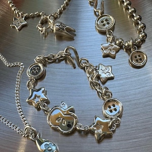 Pode incluir: Conjunto de joias prateadas com um colar com pingente de flor, uma pulseira com berloques de estrela e maçã e um colar com pingente de dente. A pulseira inclui detalhes de botões. As peças são exibidas em uma superfície metálica.